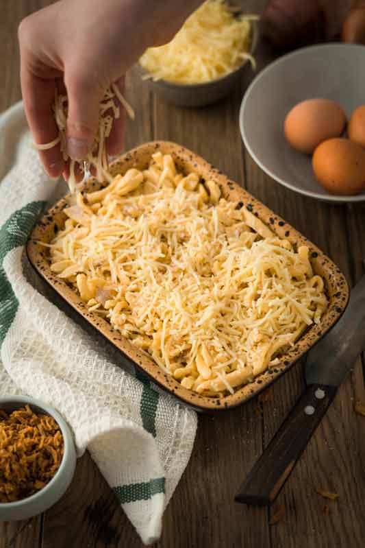 Hand streut Käse über Spätzle in einer Auflaufform auf Holzbrett, mit Schüssel und Eiern im Hintergrund.