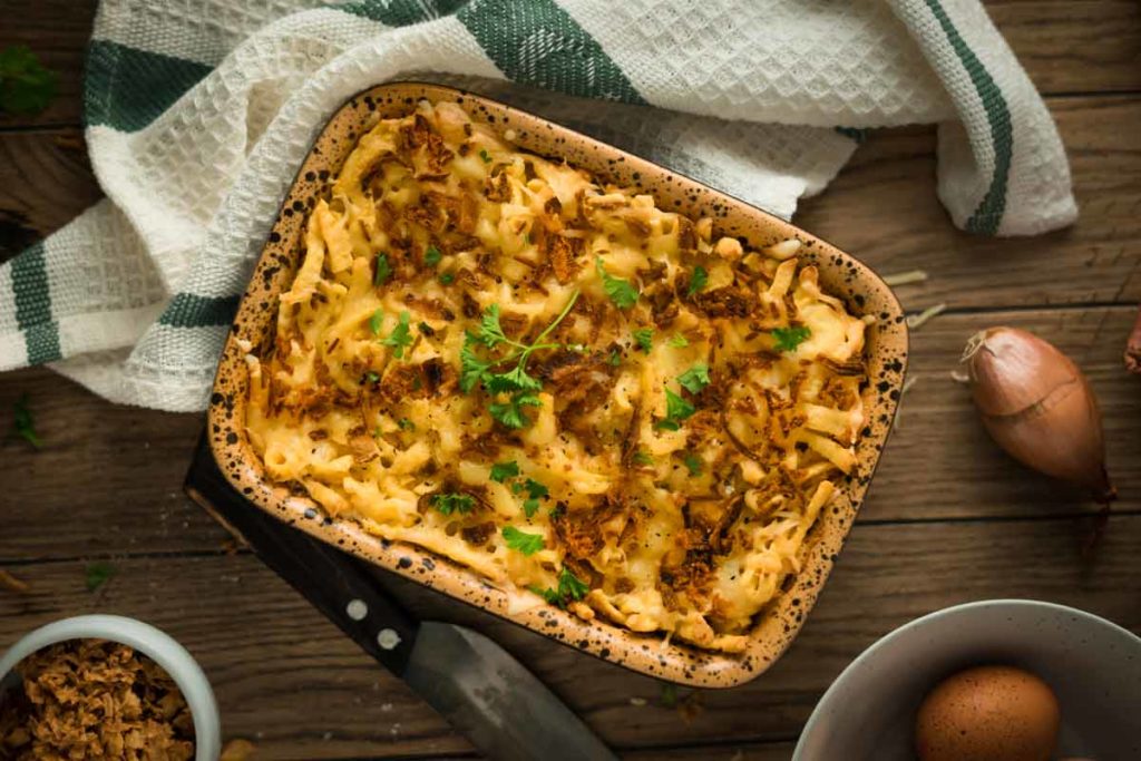 Auflauf in brauner Form mit geschmolzenem Käse, Zwiebeln und Petersilie, auf Holztisch neben einem Handtuch.
