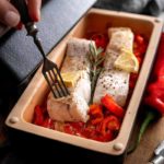Hand hält Gabel über Fischfilets mit Zitronen und Kräutern auf gerösteten Paprikastücken in einer Backform.