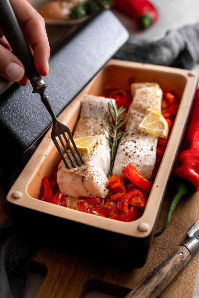 Hand hält Gabel über Fischfilets mit Zitronen und Kräutern auf gerösteten Paprikastücken in einer Backform.