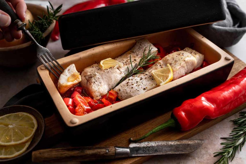 Zwei Fischfilets mit Zitrone und Rosmarin in einer Backform, umgeben von Paprika und Zitronenscheiben.