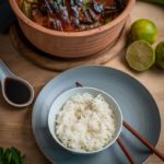 Keramiktopf mit gebratenem Fleisch und Gemüse, daneben eine Schale Reis mit Stäbchen, Sojasauce, Limetten und Petersilie.