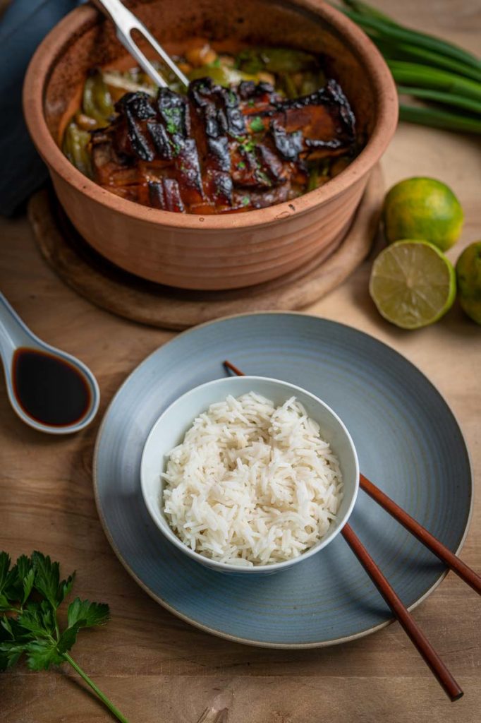 Keramiktopf mit gebratenem Fleisch und Gemüse, daneben eine Schale Reis mit Stäbchen, Sojasauce, Limetten und Petersilie.