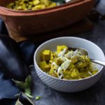 Weiße Schüssel mit gelbem Curry aus Tofu und Lauchzwiebeln, im Hintergrund eine Terrakotta-Form und eine Aubergine.