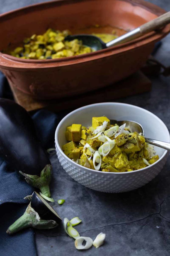 Weiße Schüssel mit gelbem Curry aus Tofu und Lauchzwiebeln, im Hintergrund eine Terrakotta-Form und eine Aubergine.