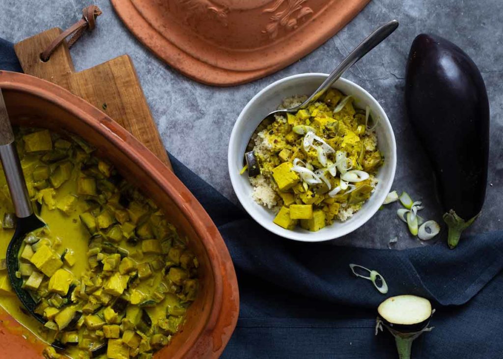 Terracotta-Topf mit Curry-Gemüse, serviert in einer Schüssel mit Couscous und Frühlingszwiebeln. Daneben eine frische Aubergine.