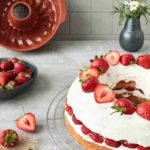 Erdbeertorte mit Sahne, dekoriert mit Erdbeeren, auf einem Gitter. Gugelhupfform und Vase im Hintergrund.