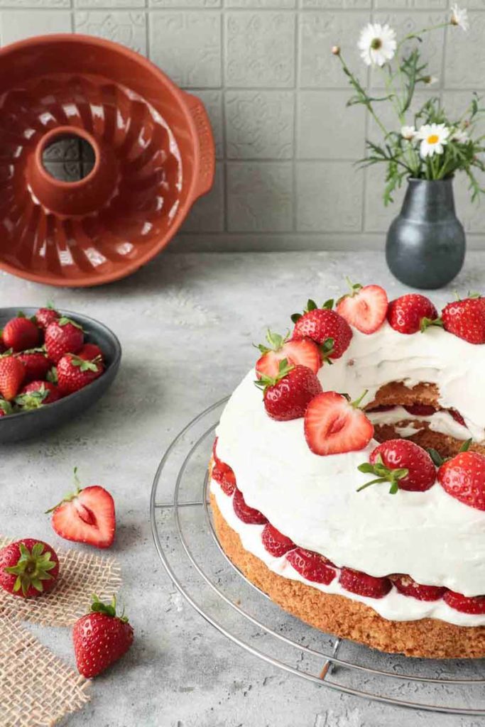 Erdbeertorte mit Sahne, dekoriert mit Erdbeeren, auf einem Gitter. Gugelhupfform und Vase im Hintergrund.