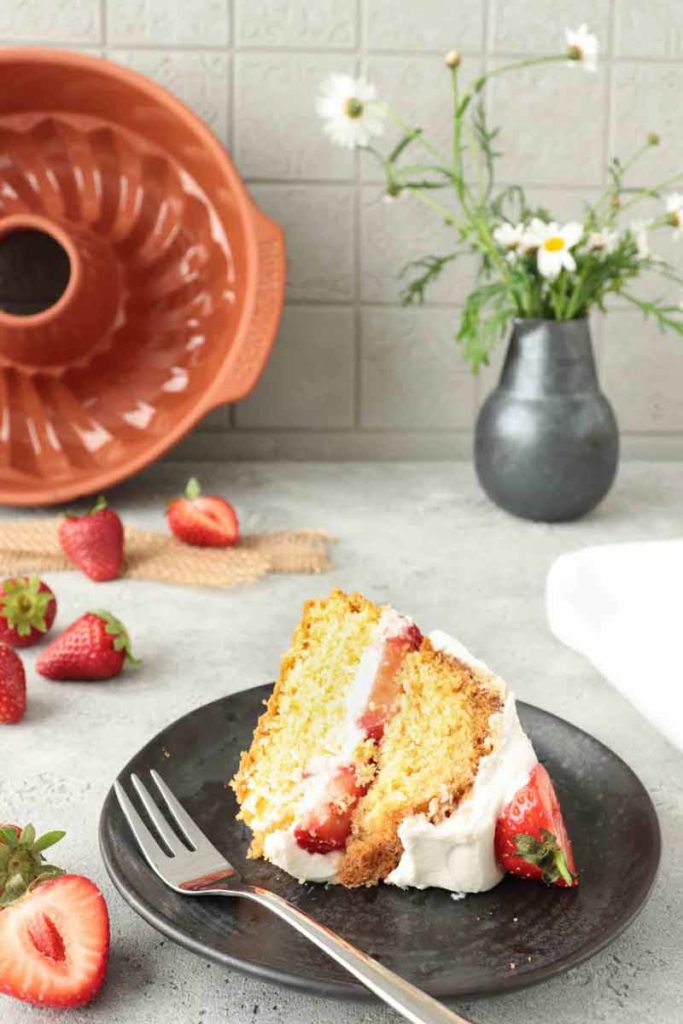 Ein Stück Erdbeerkuchen auf schwarzem Teller mit Gabel, umgeben von frischen Erdbeeren, Vase im Hintergrund.