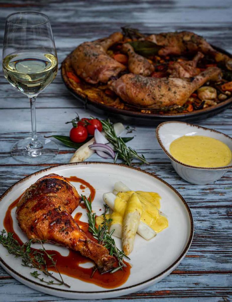 Gegrilltes Hähnchen mit Spargel und Sauce, neben einem Glas Weißwein und tomatendekoriertem Rosmarin.