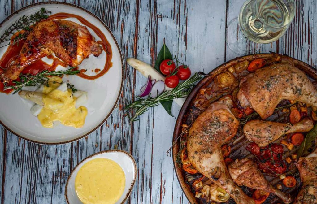 Gebackene Hähnchenschenkel mit Gemüse, Spargel mit Sauce und einem Glas Weißwein auf rustikalem Holztisch.