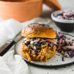 Burger mit Pulled Pork und Krautsalat auf einem Teller, daneben ein Tontopf und eine Schüssel Krautsalat.