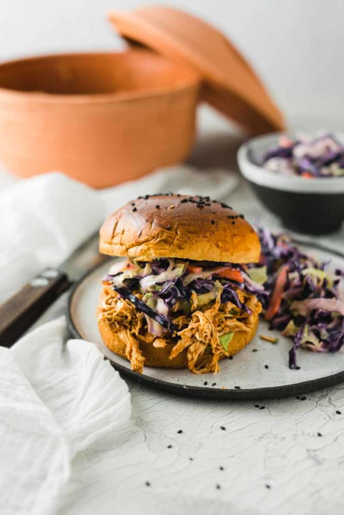 Burger mit Pulled Pork und Krautsalat auf einem Teller, daneben ein Tontopf und eine Schüssel Krautsalat.