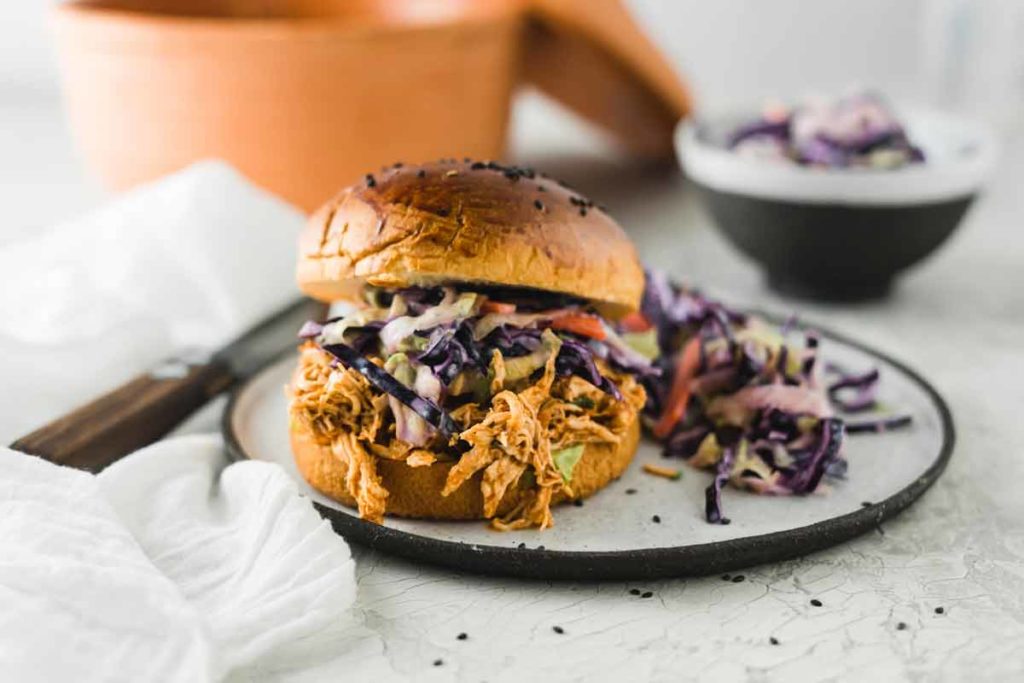 Brötchen mit gezupftem Hühnchen und Krautsalat auf Teller, im Hintergrund Salatschüssel.