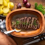 Bräter mit geschnittenem Braten, daneben Kartoffeln, rote Zwiebeln und Petersilie auf Holztisch.