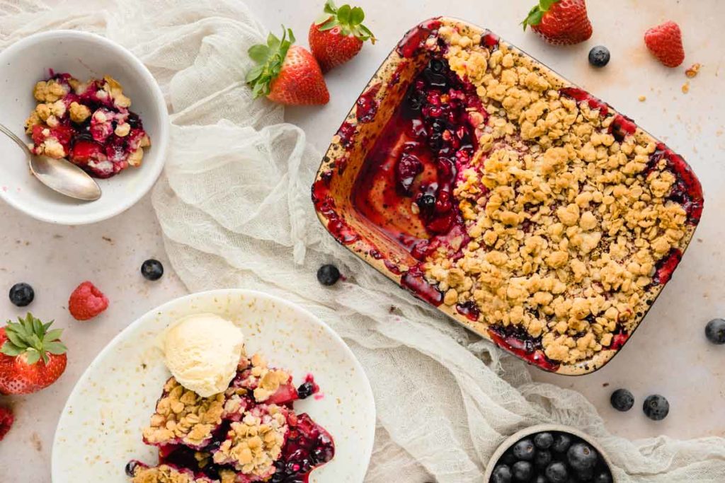 Beeren-Crumble mit Streuseln in einer Backform, serviert mit Vanilleeis, umgeben von frischen Beeren.