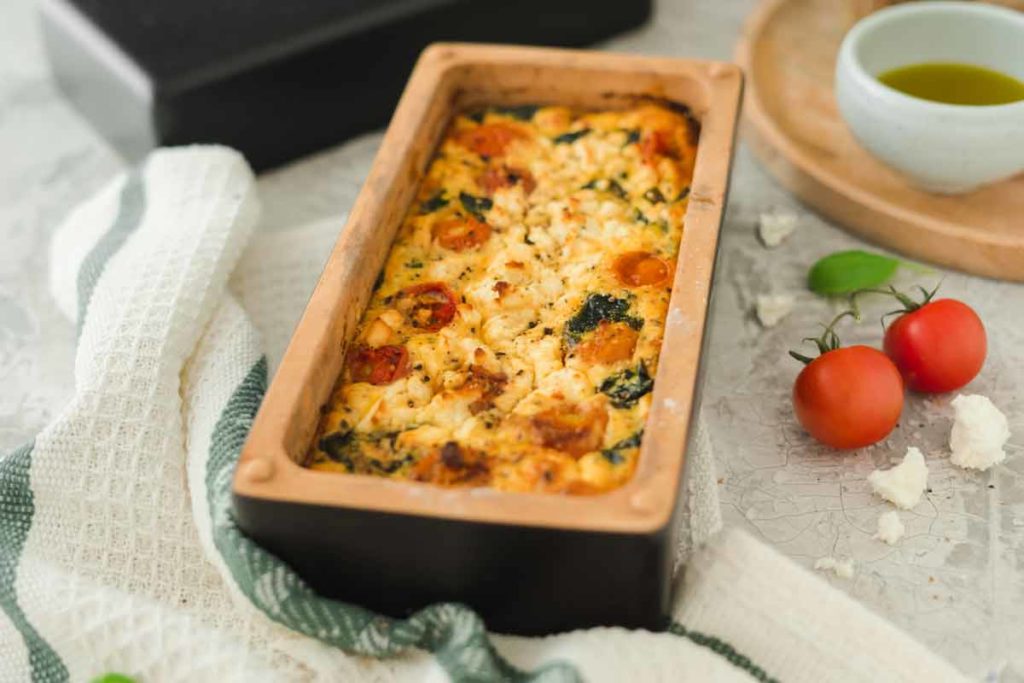 Auflaufform mit gebackenem Feta, Kirschtomaten und Spinat. Daneben liegen frische Tomaten und eine Schale mit Öl.