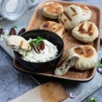 Ein Tablett mit frischen Fladenbroten und einer Schale mit Frischkäse-Dip, garniert mit Datteln und Kräutern.
