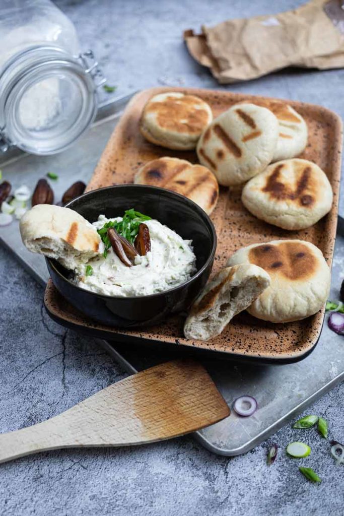 Ein Tablett mit frischen Fladenbroten und einer Schale mit Frischkäse-Dip, garniert mit Datteln und Kräutern.