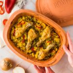 Hähnchenkeulen auf Reis mit Paprika in einer Terrakotta-Form, neben Zwiebel und Paprikascheiben auf einem Tisch.