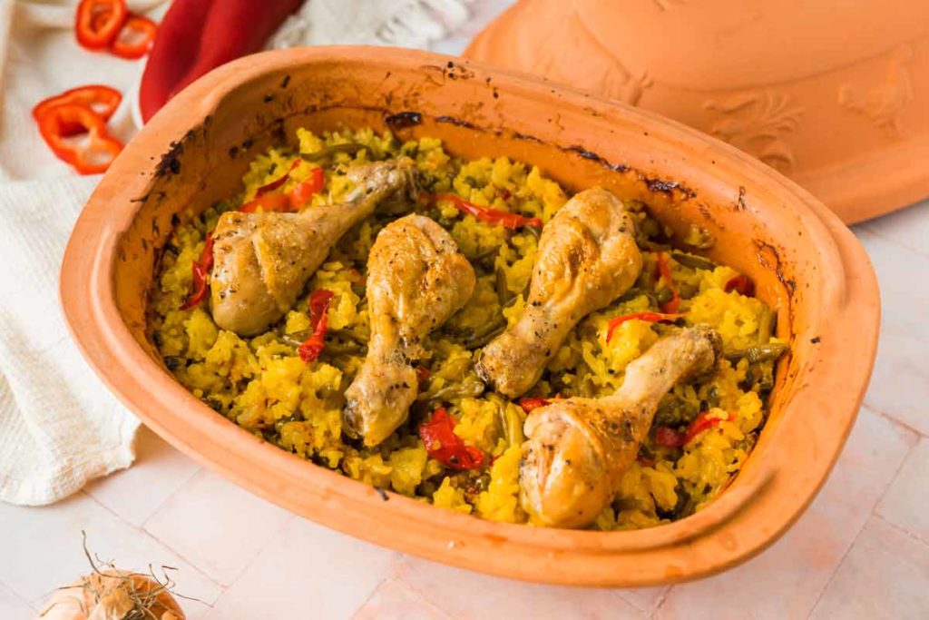Hühnchenschenkel auf gelbem Reis mit Paprika in einer Terrakotta-Form, auf einem hellen Tisch.