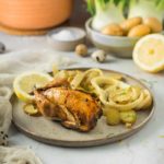 Geröstetes Hühnchen auf Teller mit Zitronenscheiben und Fenchel, im Hintergrund Gemüse und Geschirr.