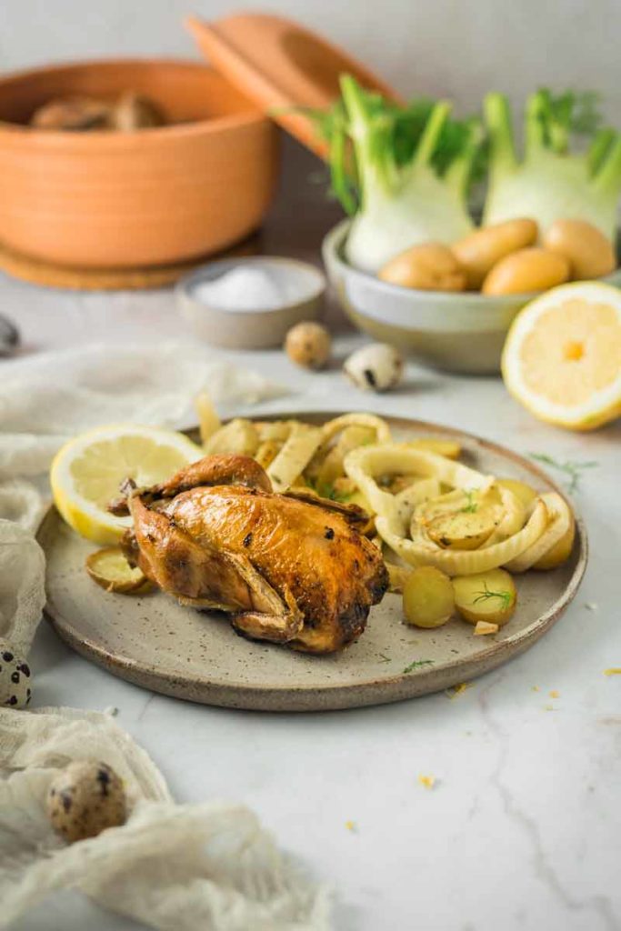 Geröstetes Hühnchen auf Teller mit Zitronenscheiben und Fenchel, im Hintergrund Gemüse und Geschirr.