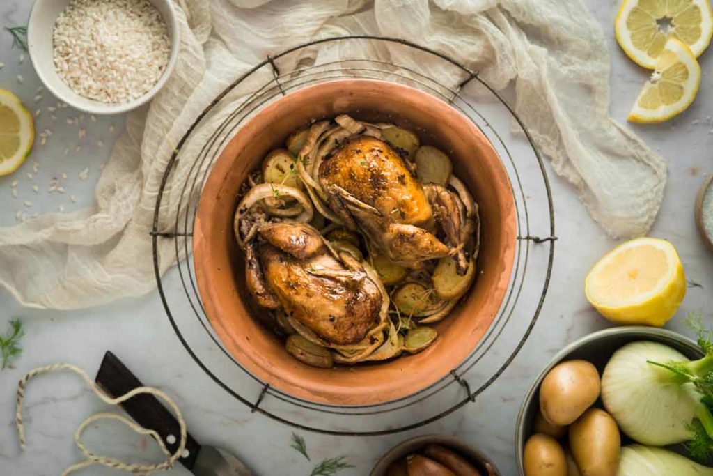 Zwei gebratene Wachteln mit Kartoffeln und Zwiebeln in einem Tontopf, umgeben von Zitronenscheiben, Reis und Kartoffeln.