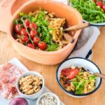 Schüssel mit Nudelsalat, Rucola und Tomaten, daneben Walnüsse, Zwiebeln und Salami auf einem Holzbrett.