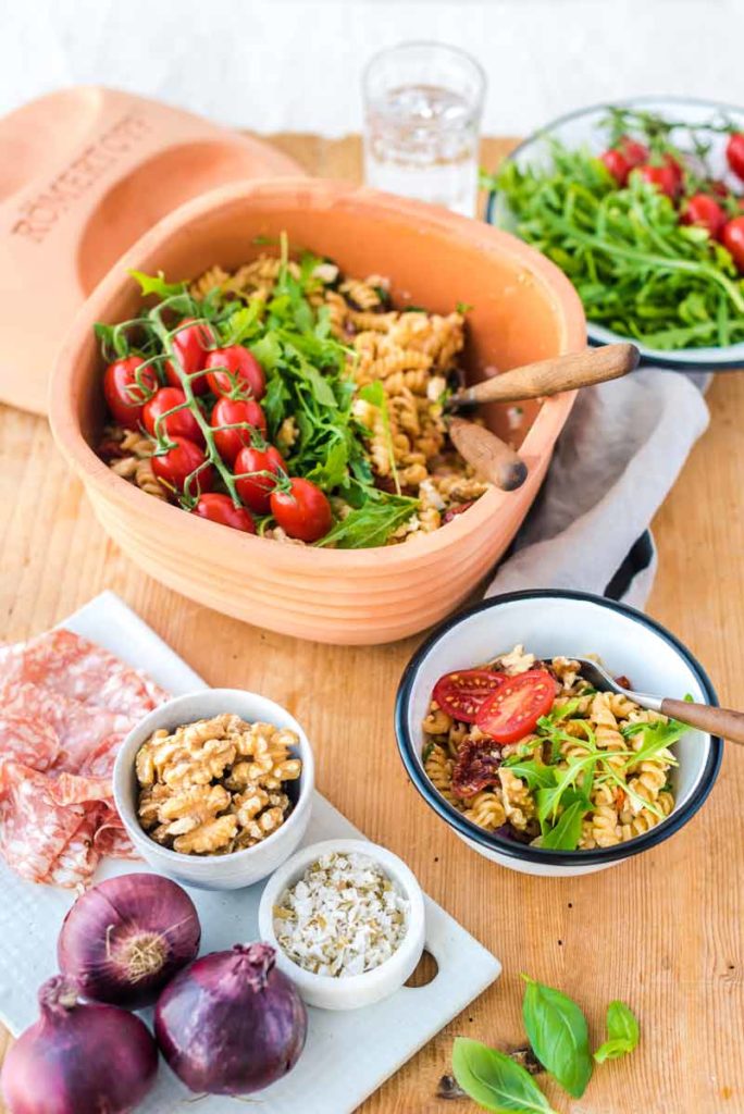 Schüssel mit Nudelsalat, Rucola und Tomaten, daneben Walnüsse, Zwiebeln und Salami auf einem Holzbrett.