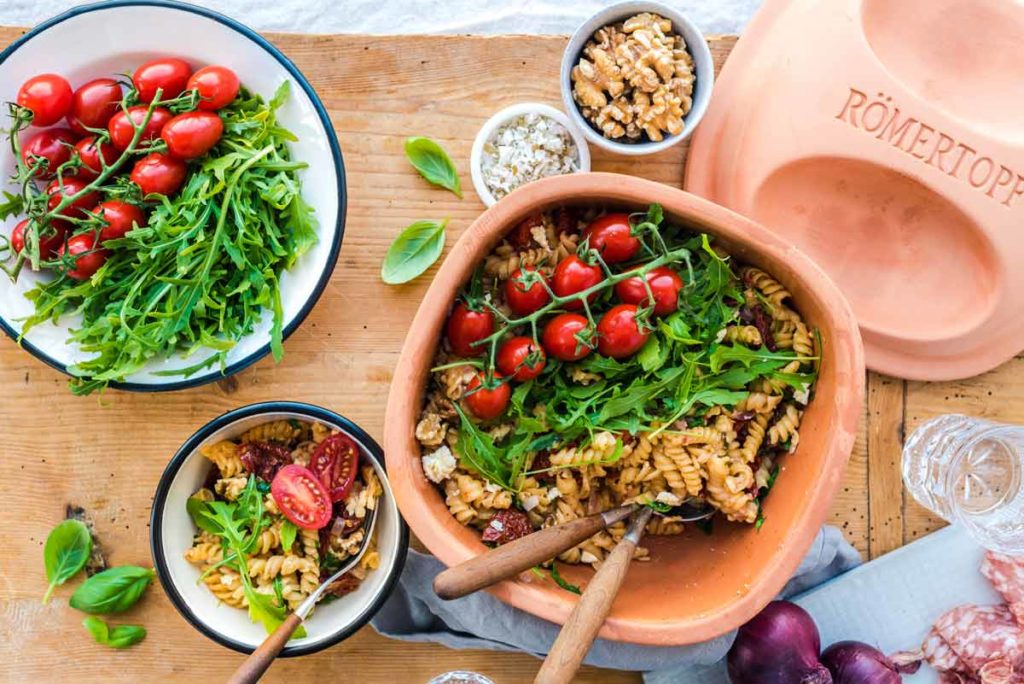 Pasta mit Rucola, Kirschtomaten und Walnüssen in Römertopf, serviert auf einem Holztisch mit frischen Zutaten und Salami.