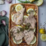 Gebratenes Fleisch mit Knoblauch, Zitronenscheiben und Rosmarin auf einem Blech, umgeben von frischen Zutaten.