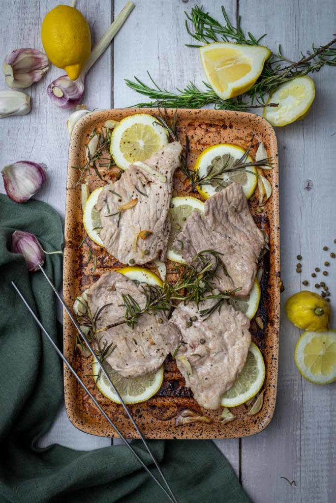 Gebratenes Fleisch mit Knoblauch, Zitronenscheiben und Rosmarin auf einem Blech, umgeben von frischen Zutaten.