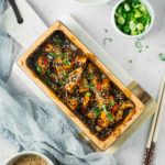 Eine servierte Schale mit gebackener Aubergine in Soße, garniert mit Sesam und Frühlingszwiebeln. Daneben Reis und Sesamsamen.