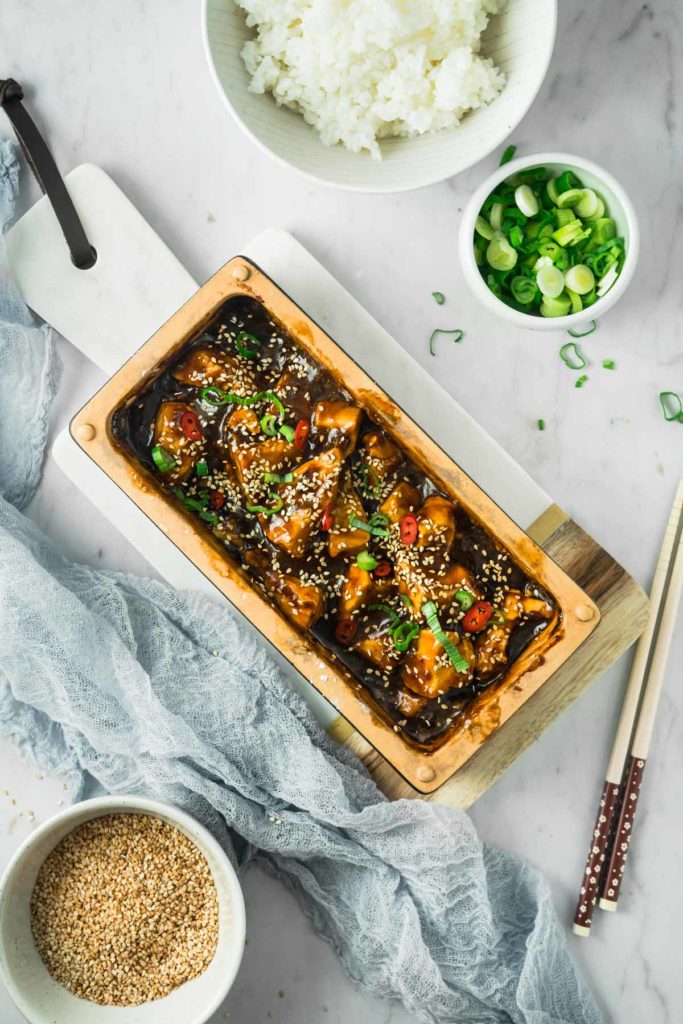 Eine servierte Schale mit gebackener Aubergine in Soße, garniert mit Sesam und Frühlingszwiebeln. Daneben Reis und Sesamsamen.