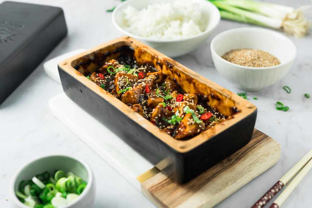 Essen in schwarzem Behälter mit Sesam, Reis und Frühlingszwiebeln, daneben Schale mit begleitenden Zutaten auf Tisch.