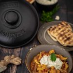 Schüssel mit Blumenkohl-Curry und Joghurt, umgeben von Naan-Brot, Reis und einem Römertopf auf Holztisch.