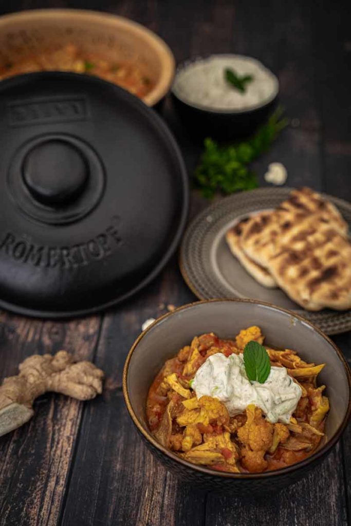 Schüssel mit Blumenkohl-Curry und Joghurt, umgeben von Naan-Brot, Reis und einem Römertopf auf Holztisch.