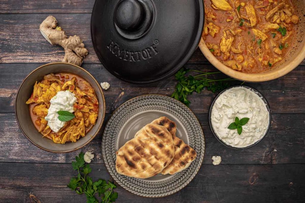 Indisches Curry mit Hühnchen, Joghurt-Dip, Naan-Brot und frischem Ingwer auf einem dunklen Holztisch.