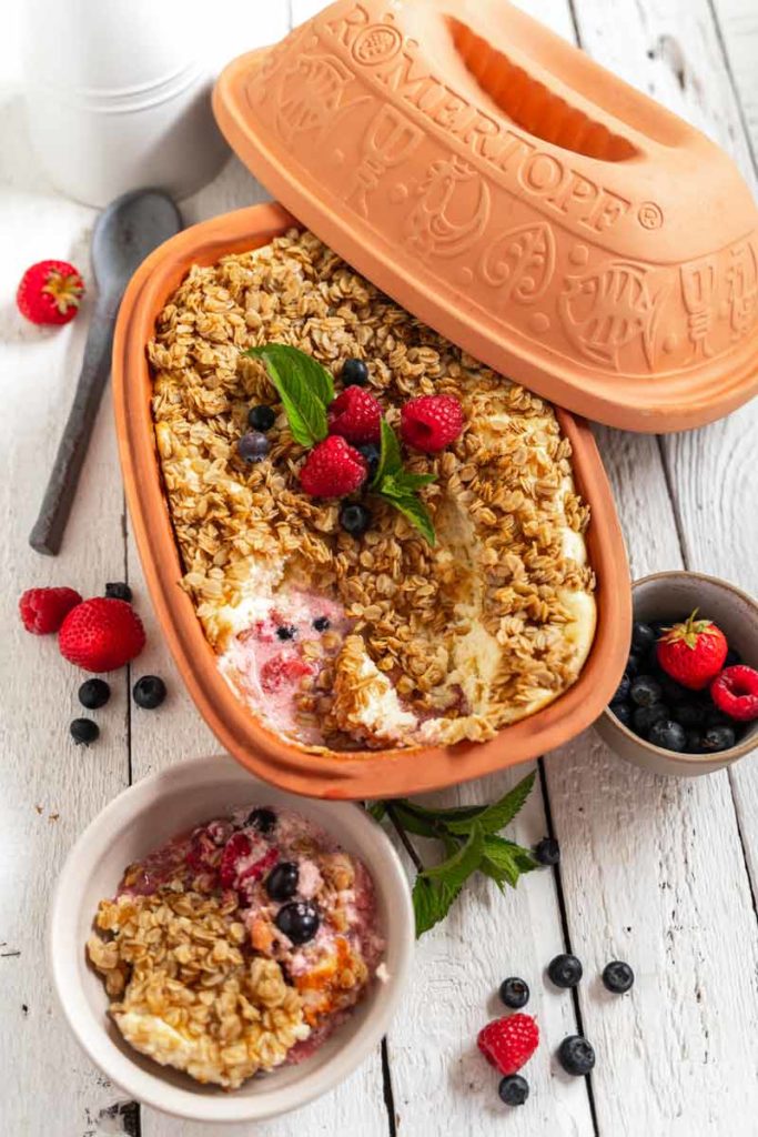 Lehm-Backform mit Haferauflauf und Beeren, darunter eine Portion in einer Schale. Deko mit frischen Beeren und Minze.