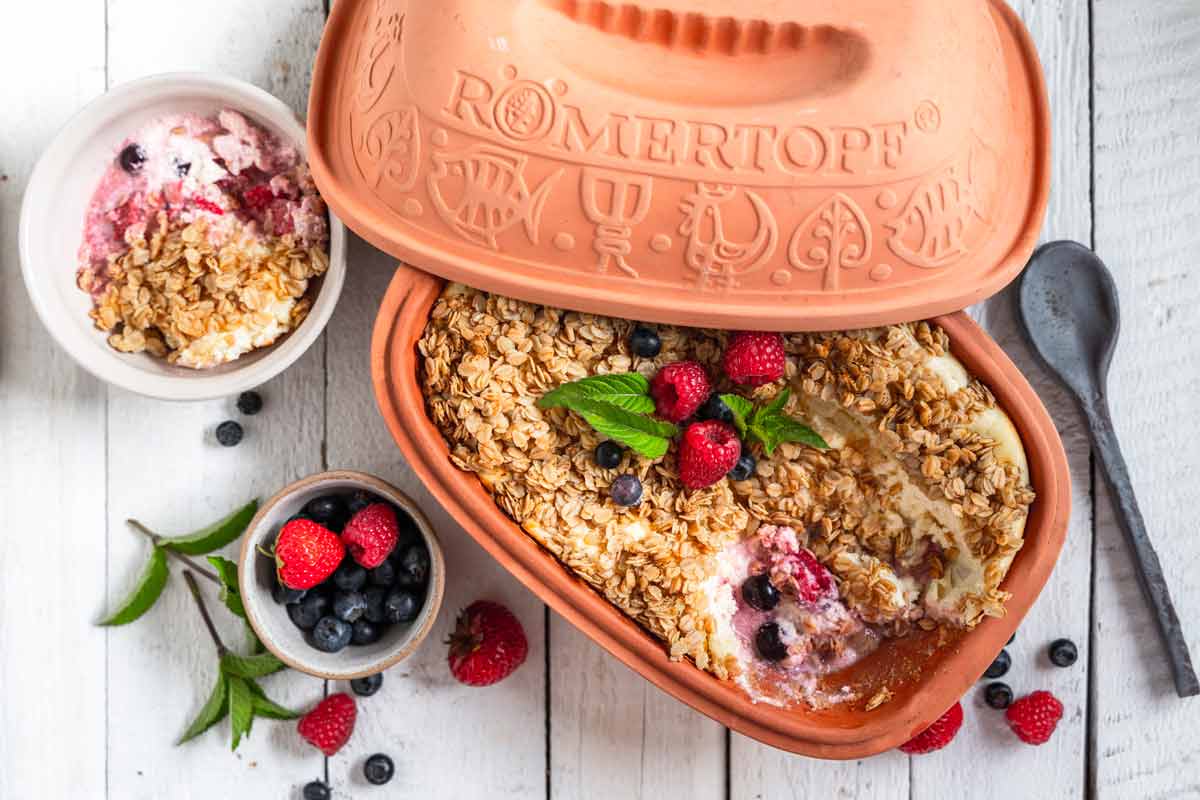 Römertopf mit Haferauflauf, Beeren und Minze, daneben Schüssel mit Dessert und frischen Beeren auf hellen Holzboden.