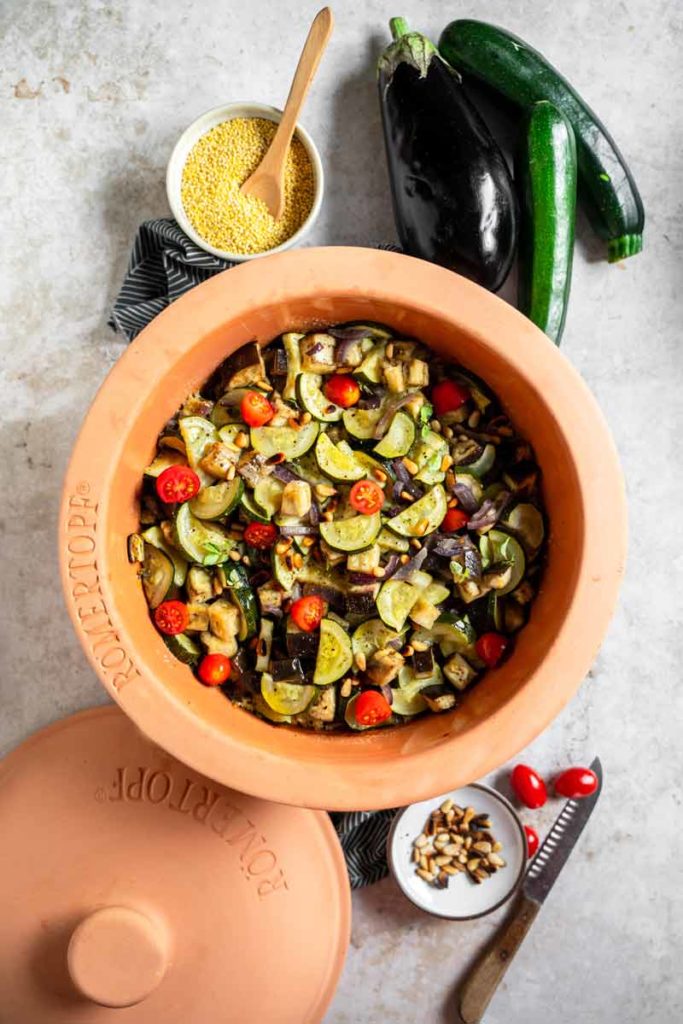 Gemüse-Ratatouille in einem Römertopf mit Zucchini, Auberginen, Tomaten und Pinienkernen, daneben Hirse und ganze Gemüsesorten.