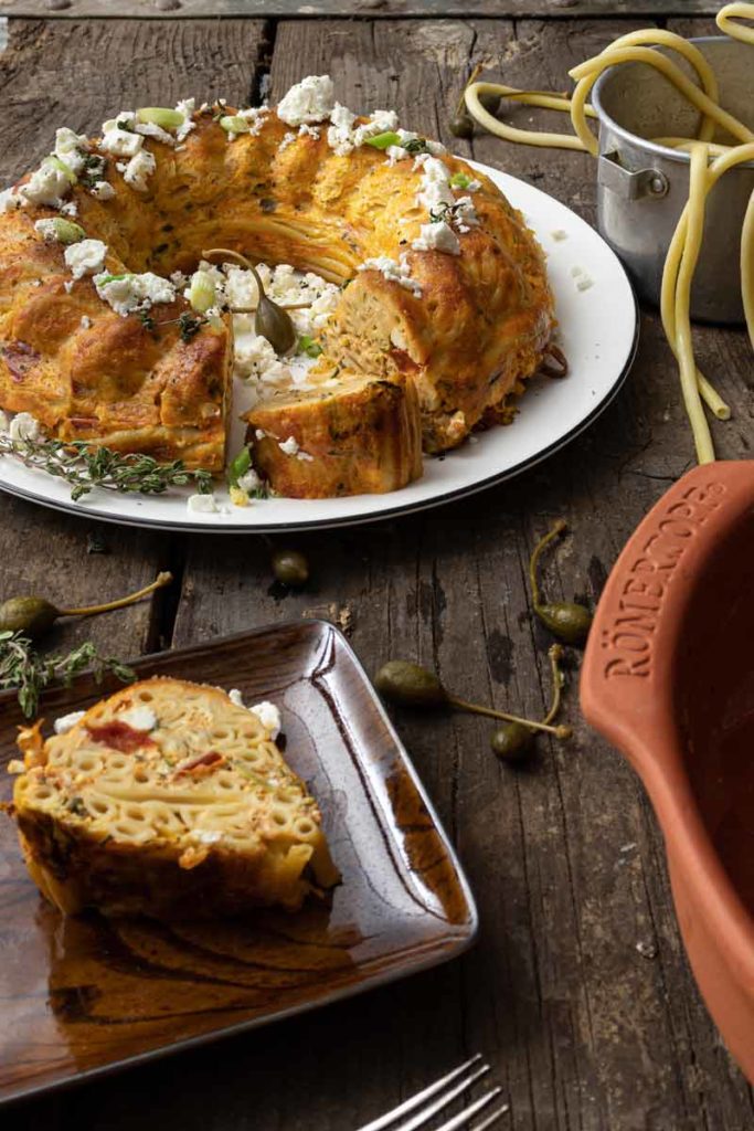 Gugelhupf aus Nudeln auf Teller mit Feta und Kräutern, ein Stück auf braunem Teller, Holztisch, Topf im Hintergrund.