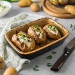 Gebackene Kartoffeln mit Sauerrahm und Schnittlauch in einer Keramikform, daneben frische Kartoffeln und Frühlingszwiebeln.