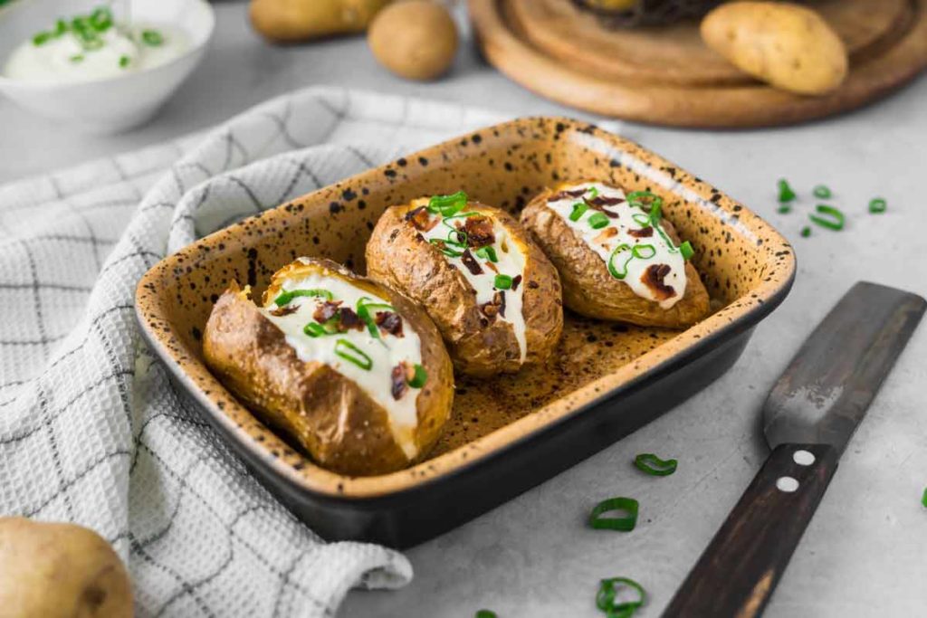 Drei Ofenkartoffeln mit Sauerrahm und Frühlingszwiebeln in einer Backform, daneben ein Messer und ein Geschirrtuch.
