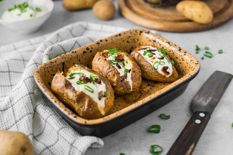 Drei Ofenkartoffeln mit Sauerrahm und Frühlingszwiebeln in einer Backform, daneben ein Messer und ein Geschirrtuch.