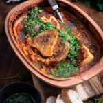 Osso Buco in rustikaler Auflaufform mit Gremolata, serviert mit Baguettescheiben, Knoblauch und Zitronenhälften im Hintergrund.