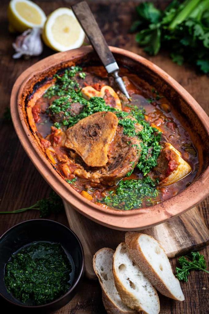 Osso Buco in rustikaler Auflaufform mit Gremolata, serviert mit Baguettescheiben, Knoblauch und Zitronenhälften im Hintergrund.