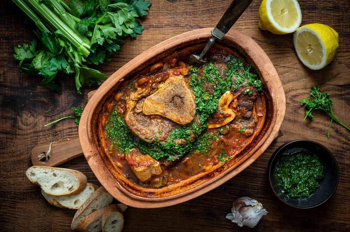 Ossobuco in einer Keramikschale mit Kräutern garniert, daneben Baguette, Zitrone und Kräuter auf Holzbrett.