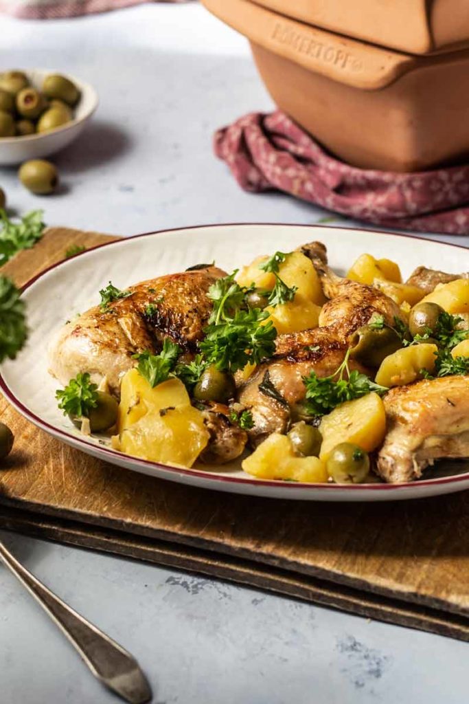 Gebratenes Hähnchen mit Kartoffeln, Oliven und Petersilie auf einem Teller vor einem Römertopf.